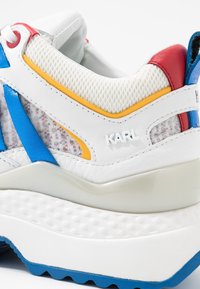 Vit, blå, röd och gul sneakersklack med meshtyg, tjock texturerad sula och "KARL" logotyp på sidan.