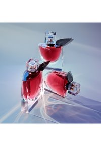 Trois flacons de parfum en verre transparent contenant un liquide rouge profond et ornés d'accents en tissu noir, disposés sur une surface réfléchissante avec un éclairage doux.