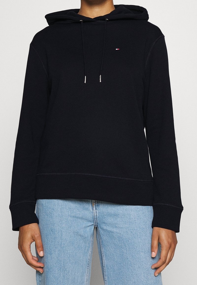 Sweat à capuche noir avec cordons et petit logo rouge, blanc et bleu sur la poitrine, porté avec un jean bleu clair.