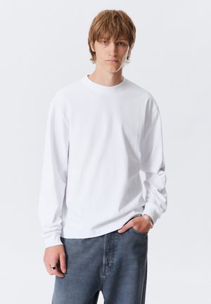 Ung mand med lysebrunt hår har en almindelig hvid langærmet skjorte, blå jeans, sølvarmbånd og ringe på, og står imod en hvid baggrund.