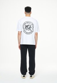 Bílé bavlněné tričko s kulatou černou grafikou na zádech včetně textu. Kombinováno s černými kalhotami rovného střihu a světlými teniskami.