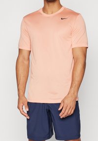 Světle růžové sportovní tričko s krátkými rukávy a kulatým výstřihem. Na hrudi je černé logo Nike. Měkká, hladká textilie.