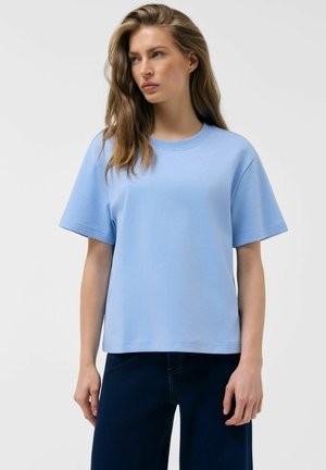 Kobieta o długich włosach, ubrana w jasnoniebieski T-shirt z krótkim rękawem i ciemnoniebieskie spodnie, stoi na tle jednolitego jasnego tła.