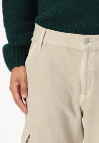 Pantalons cargo en coton beige avec une texture lisse, présentant une poche frontale, une fermeture à bouton et une coupe légèrement décontractée.