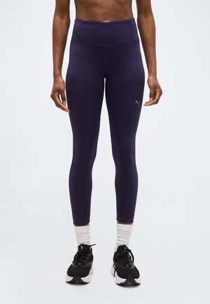 Άτομο που φοράει ψηλόμεσα σκουρόμωβ κολάν, λευκές κάλτσες και αθλητικά παπούτσια ασπρόμαυρα, στέκεται μπροστά σε απλό φόντο.