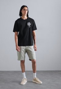 Schwarzes T-Shirt mit kurzen Ärmeln und weißem Graf design, kombiniert mit hellgrünen Shorts und beigen Sneakers. Weiße Socken sichtbar.