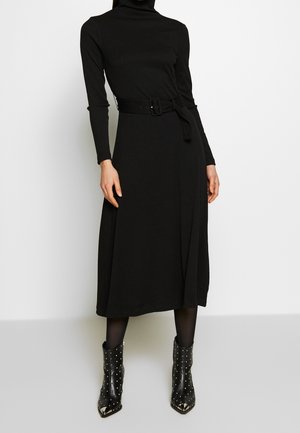 Kvinde iført en sort langærmet midi-kjole med bælte, kombineret med sorte ankelstøvler med nitter og sorte strømpebukser foran en ensfarvet baggrund.