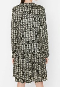 Långärmad klänning med ett blommönster i svart på en ljus olivbakgrund, med en rynkad fåll och mjukt, texturerat tyg.