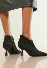 Botas de tobillo de ante negro con punta afilada y cremallera lateral. Presentan un tacón delgado y angulado y una textura suave y mate.