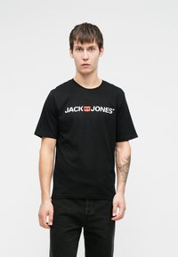 Melna kokvilnas T-kreklu ar "JACK & JONES" logotipu baltā un sarkanā krāsā. Īsām piedurknēm, apaļa kakla izgriezuma, valkāts kopā ar tumšiem džinsiem.