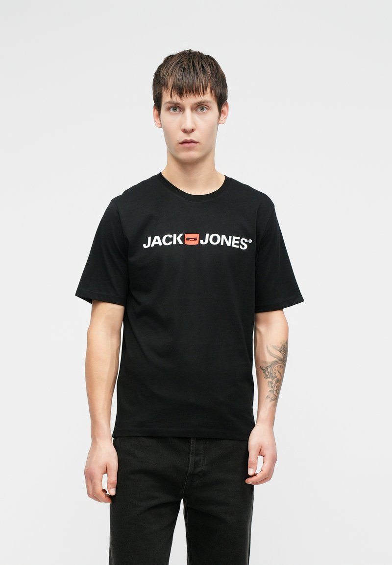 Melna kokvilnas T-kreklu ar "JACK & JONES" logotipu baltā un sarkanā krāsā. Īsām piedurknēm, apaļa kakla izgriezuma, valkāts kopā ar tumšiem džinsiem.