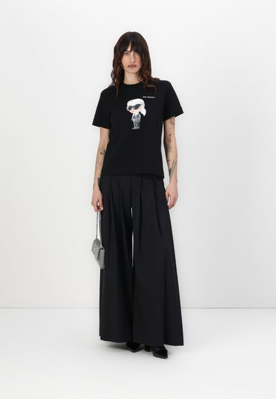 T-shirt en coton noir avec un graphique de personnage de bande dessinée, associé à un pantalon large noir et un sac à main argenté. Texture lisse, design décontracté.