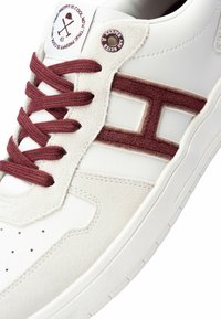 Białe skórzane sneakersy z burgundowymi zamszowymi akcentami i sznurówkami. Posiadają teksturowaną podeszwę środkową oraz geometryczny wzór na boku.