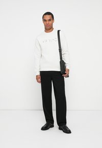 Sudadera blanca con el logo en relieve "GUESS", combinada con pantalones negros y zapatos negros. Un bolso cruzado negro aporta contraste.