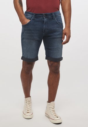 Man draagt donkerblauwe opgerolde denim shorts, bordeauxrode shirt en witte hoge sneakers, staande met handen in de zakken.