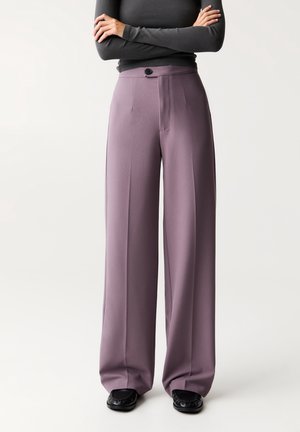Personne portant un pantalon mauve taille haute à jambes larges avec un pli à l'avant, une fermeture à boutons noirs, des chaussures noires et un haut ajusté à manches longues couleur charbon.