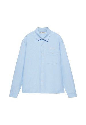 Jeune homme portant une chemise bleu clair à boutons, un jean ample clair et des chaussures noires, debout devant un fond clair uni.