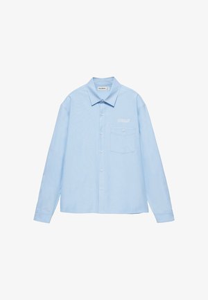Giovane uomo con camicia azzurro chiaro abbottonata, jeans larghi chiari e scarpe nere, in piedi davanti a uno sfondo chiaro e uniforme.