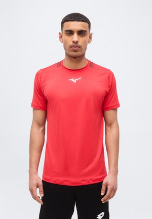Mannelijk model met kort zwart haar, gekleed in een rood sportshirt en zwarte korte broek, staand tegen een effen lichte achtergrond.