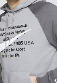 Grå hoodie med en stor vit Nike swoosh-logga, svart text och kontrasterande mörkgrå ärmar. Designad för sport med en känguruficka.