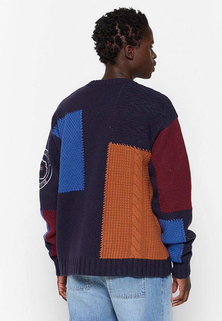 Suéter de punto con un diseño de patchwork en azul marino, azul, burdeos y naranja; presenta patrones texturizados, puños acanalados y un cuello redondo.