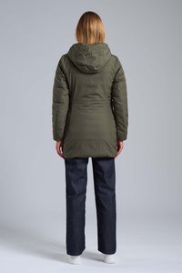Persona con capelli castano chiaro di lunghezza media indossa una giacca a giacca verde oliva, jeans scuri e scarpe bianche, in piedi con la schiena rivolta.