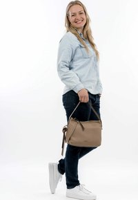 Borsa a tracolla color tan con superficie testurizzata e una lunga tracolla regolabile, abbinata a jeans scuri e sneakers bianche. Il modello si trova in piedi su uno sfondo bianco semplice.