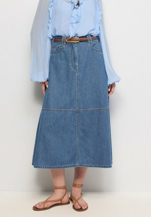 Mujer con una blusa azul claro con volantes metida dentro de una falda vaquera de largo midi con un cinturón marrón y sandalias marrones con tira en el tobillo.