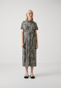 Collared, short-sleeve dress in a paisley print with shades of green, navy, and beige; belted waist; paired with black and white sandals.