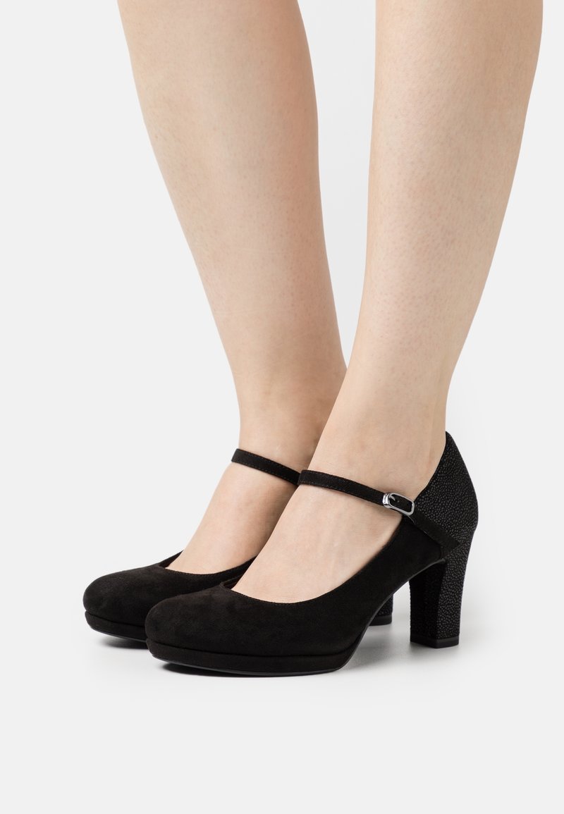 Pieds portant des talons Mary Jane en daim noir avec brides à la cheville et talons blocs texturés sur fond blanc.