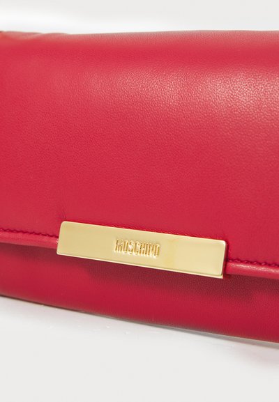 Rdeča usnjena denarnica z gladko teksturo, ki ima zlato kovinsko okrasje z imenom blagovne znamke "Moschino."