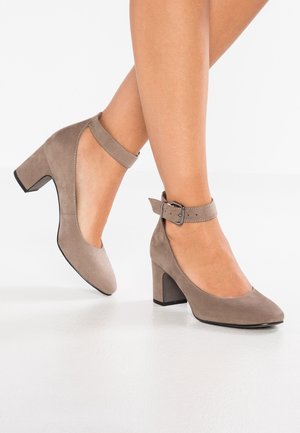 Beżowe zamszowe szpilki z okrągłym noskiem i paseczkiem na kostce. Posiadają blokowy obcas i kontrastującą czarną podeszwę. Gładka tekstura.