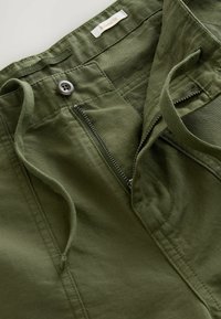 Relaxed fit olive green pants with a zipper fly and button closure. Features a textured fabric with stitching accents and a label inside.