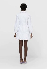 Robe en coton blanc avec une taille cintrée, des manches longues bouffantes et un ourlet à volants. Portée avec des talons lavande, vue de dos.