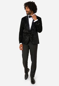 Moški nastavlja črno metuljčkasto kravato, oblečen v bleščečo smoking jakno, belo srajco, črne hlače in polirane črne večerne čevlje proti beli ozadju.