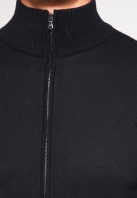 Svart ribbad zip-sweater med hög krage, metallisk dragkedja och subtil logotypdetalj på bröstet. Texturerad tygdesign.