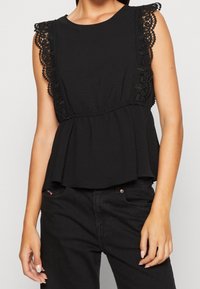 Top negro peplum con adornos de encaje en los hombros, escote redondo y tela texturizada. Combinado con vaqueros negros, que presentan un ligero vuelo en el dobladillo.