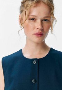 Robe sans manches bleu marine avec un col rond et trois boutons noirs sur le devant. Texture de tissu lisse et design minimaliste.