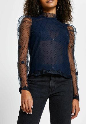 Kobieta ubrana w przezroczystą granatową bluzkę z długim rękawem w groszki, z falbanianymi mankietami i wysokim kołnierzem, w połączeniu z czarnymi dżinsami.