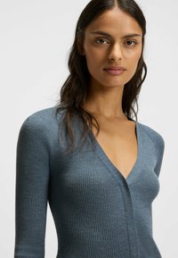 Ribbad, djup V-ringad tröja i en dämpad blå tyg, med långa ärmar och en figursydd design som framhäver kroppens form.