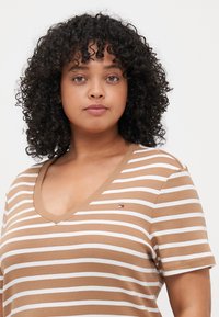 Camiseta a rayas marrón y blanca con cuello en V, mangas cortas y un pequeño detalle de logo en el pecho. Tejido de algodón suave, corte casual.