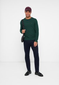 Grüner Pullover, marineblaue Hose und schwarze Schuhe. Das Model trägt eine kastanienbraune Mütze und hält eine schwarze Umhängetasche mit strukturiertem Finish.