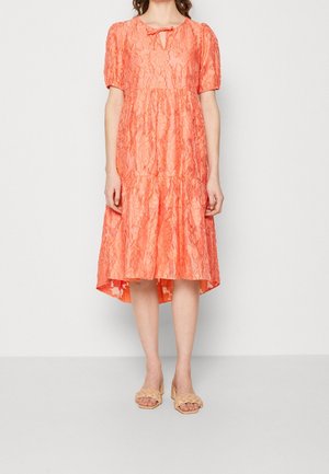 Mujer con un vestido midi de encaje coral con mangas abullonadas, escote con abertura en lágrima y sandalias beige trenzadas de tacón y dedos abiertos, de pie contra un fondo blanco.