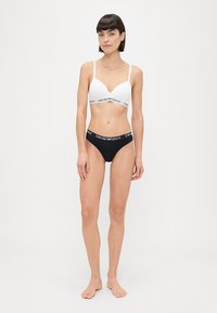 Bralet alb cu bretele reglabile și bandă elastică neagră cu logo, asociat cu chiloți negri cu talie înaltă, având bandă logo asortată.