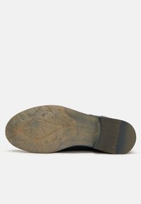 Suela de zapato de cuero negro con una superficie texturizada y desgastada, y un pequeño tacón. Patrón de relieve notable y detalles de marca grabados visibles.