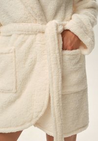 Kremowy, puszysty szlafrok z froty z wiązanym pasem, dwoma przednimi kieszeniami i miękką teksturą. Zaprojektowany z myślą o wygodzie, o długości sięgającej do połowy ud.