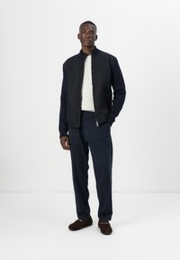 Σκούρα ναυτική βόμβα με φερμουάρ μπροστά, συνδυασμένη με ανοιχτό γκρι πλεκτό πουλόβερ και αντίστοιχες ναυτικές παντελόνες. Καφέ εσπαντρίγιες.