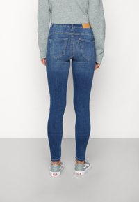 Jeans de mezclilla azul ajustados con una cintura de altura media, diseño de cinco bolsillos y parche trasero, combinados con zapatillas grises y un suéter de punto.
