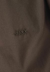 Tessuto marrone con una superficie liscia, caratterizzato dalla scritta ricamata "JUXX" in filo marrone scuro. Design semplice, senza ulteriori motivi o dettagli.