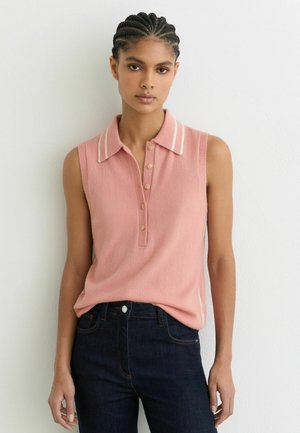 Młoda kobieta z warkoczami, ubrana w różowy bezrękawnik z kołnierzykiem i ciemnoniebieskie jeansy, stojąca na tle gładkiej białej ściany.